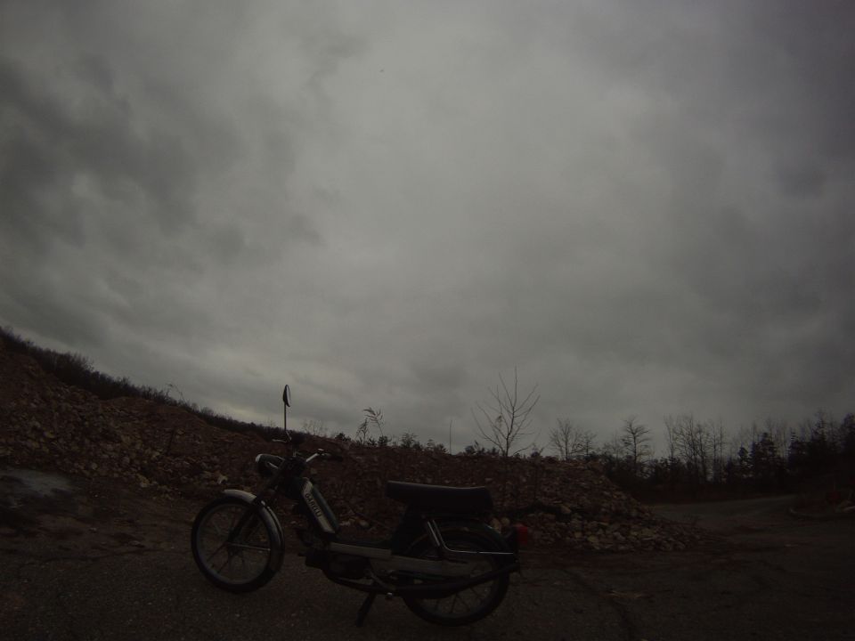 Side view of a dark moped parked on a dirt road under a cloudy sky
