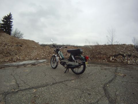 1981 Garelli Rally Sport - A green moped sits parked on a cracked asphalt surface, with large piles of rubble and debris forming the backdrop under a cloudy sky.