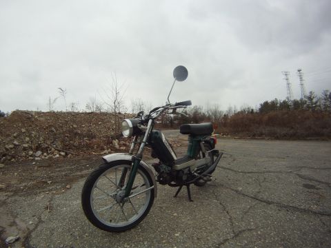 1981 Garelli Rally Sport - A green moped with a chrome fender and a round mirror is parked on an asphalt surface with a pile of rubble in the background.