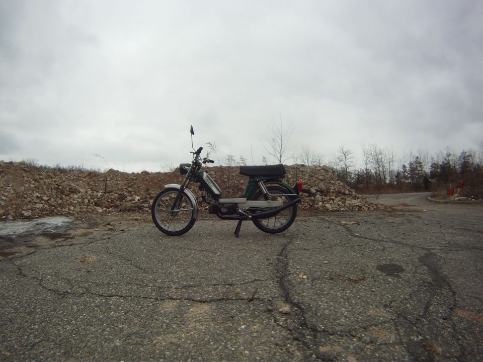 Green moped parked on cracked asphalt with piles of rocks and bare trees in the background