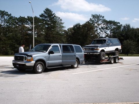 1996 Ford Bronco - A Ford Excursion tows a lifted Ford Bronco on a trailer in a parking lot.