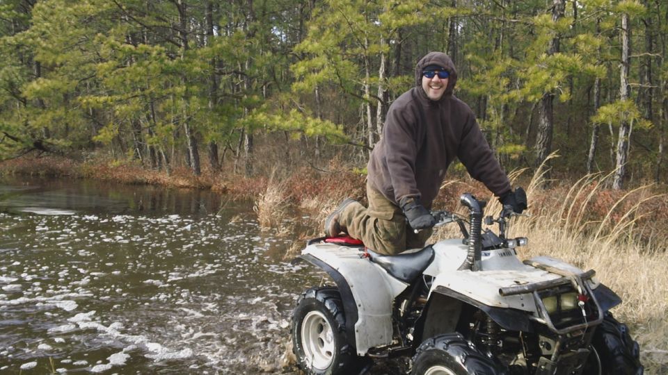 Man on white Yamaha ATV at edge of foamy pond with pine trees behind