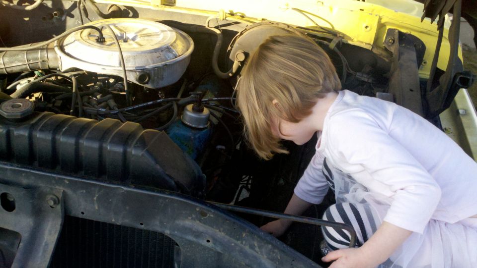 Kid in striped pants peers into engine bay of yellow vehicle