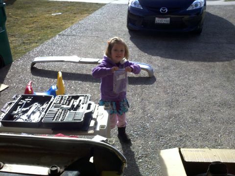 1978 Ford Bronco - A young girl stands with tools and car parts, with a black Mazda parked in the background.