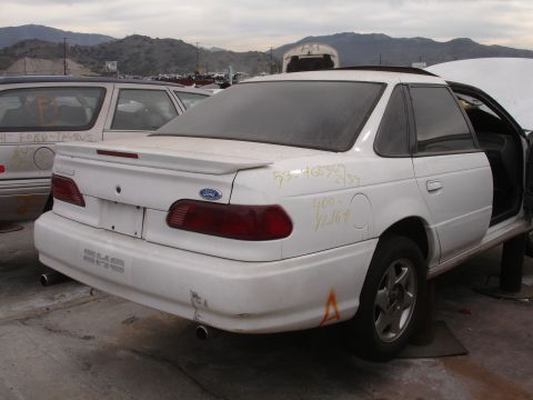 1993 Ford Taurus - A white 1993 Ford Taurus sedan sits in a junkyard, showing its rear and side profile.