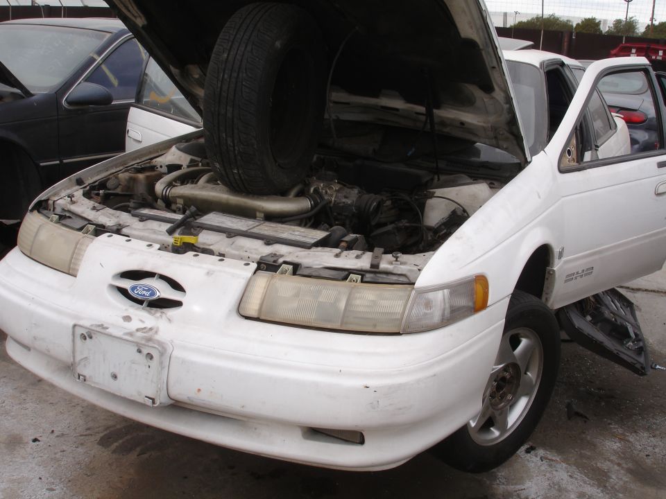 1993 Ford Taurus front view, hood open, tire in engine bay. Another car is visible behind it