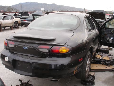 1993 Ford Taurus - A black Ford Taurus sits in a junkyard with its rear facing the camera and its driver's side door open.