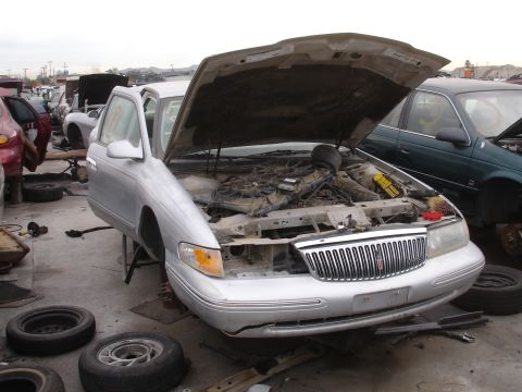1993 Ford Taurus - A silver sedan sits in a junkyard with its hood open and missing wheels.