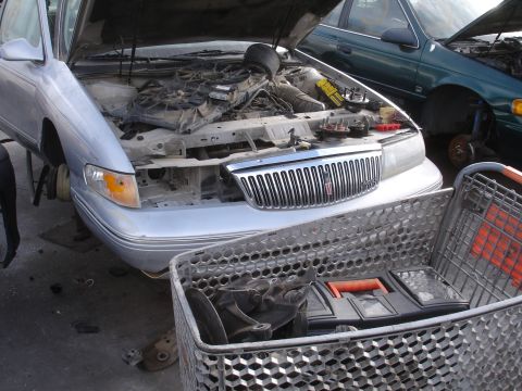 1993 Ford Taurus - A silver sedan with its hood open, revealing the engine compartment, sits next to another vehicle in a junkyard.