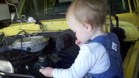 1978 Ford Bronco - A toddler in denim overalls curiously examines the engine of a yellow 1978 Ford Bronco.