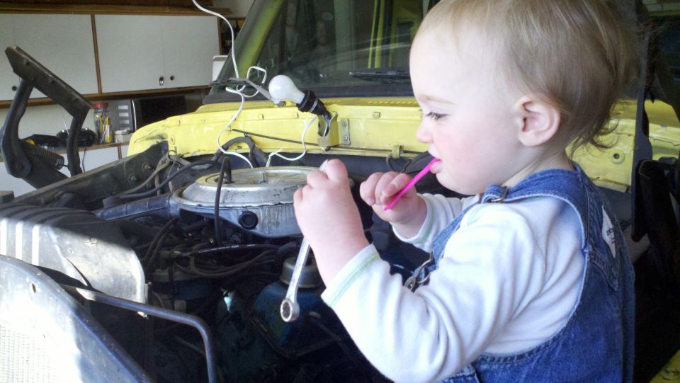 Child in denim overalls holding a wrench and toothbrush near a yellow vehicle's engine compartment