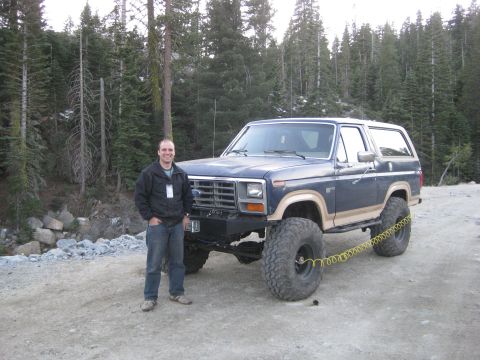 1986 Ford Bronco - A man poses with a modified Ford Bronco, equipped with oversized tires and a lifted suspension, in a wooded area.