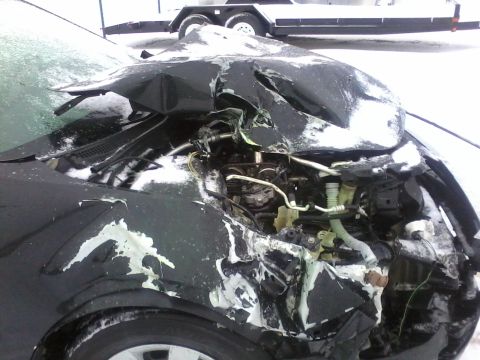 2006 Ford Fusion - The front end of a black car is heavily damaged, exposing the engine components, with snow covering the vehicle and surrounding area.