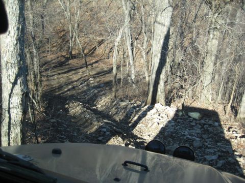 2005 Jeep Wrangler - Driving on a rough, wooded trail with rocks and trees visible.