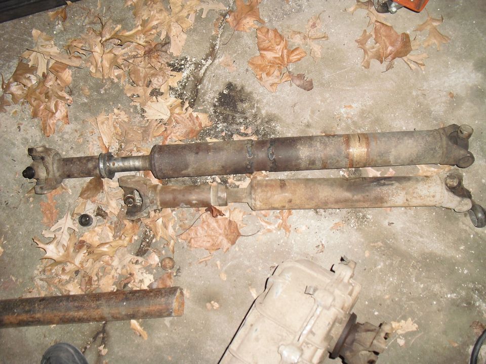 Two driveshafts and a transfer case are on the ground, covered in leaves