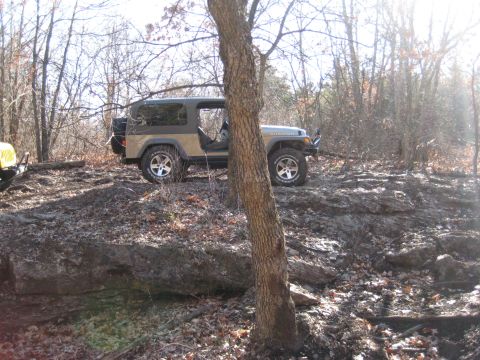 2005 Jeep Wrangler - A tan Jeep Wrangler navigates a rocky, wooded trail, with a yellow ATV visible in the background.