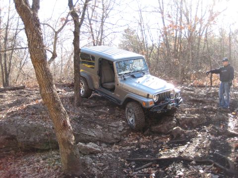 2005 Jeep Wrangler - A Jeep Wrangler is shown off-roading on a trail with trees and rocks.
