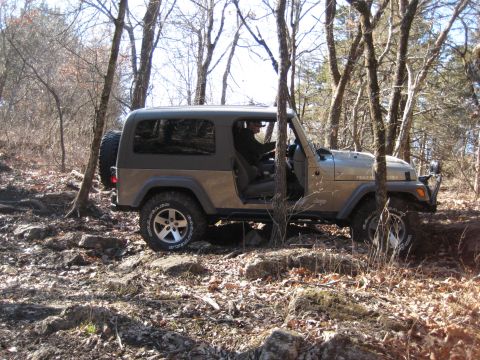 2005 Jeep Wrangler - A tan Jeep Wrangler navigates a rough, wooded path with rocks and fallen leaves.