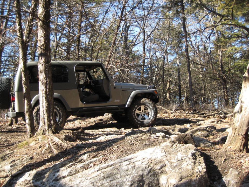 Olive green 2005 Jeep Wrangler with doors removed, parked on a rocky, wooded trail