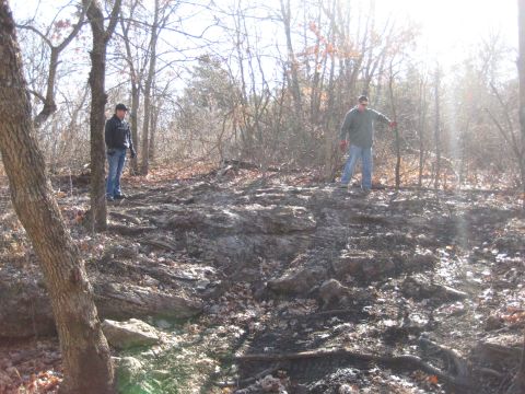2005 Jeep Wrangler - Two men are in a wooded area with rocky terrain and sparse vegetation under a bright sky.