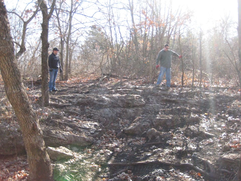 Two men stand in a sun-dappled, rocky forest clearing with bare trees and fallen leaves