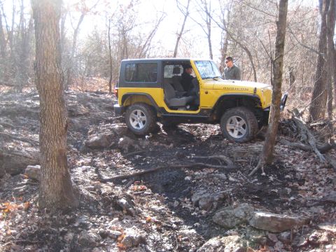 2005 Jeep Wrangler - A yellow Jeep Wrangler Rubicon is shown off-roading on a rough, wooded terrain with a person standing outside the vehicle.