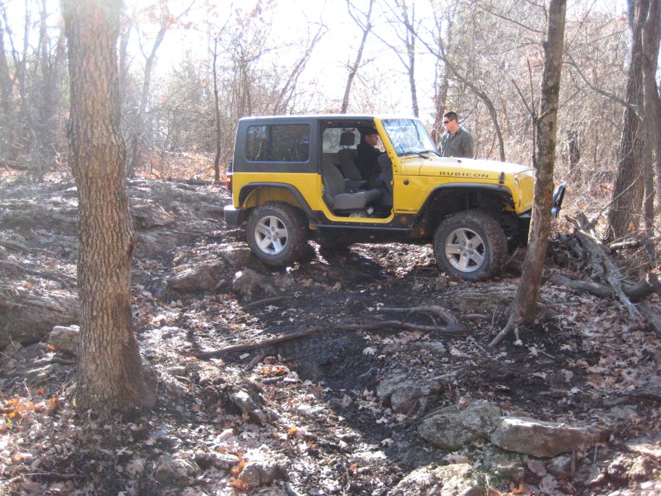 Yellow 2005 Jeep Wrangler Rubicon navigating a rocky, wooded trail. Driver and passenger visible inside