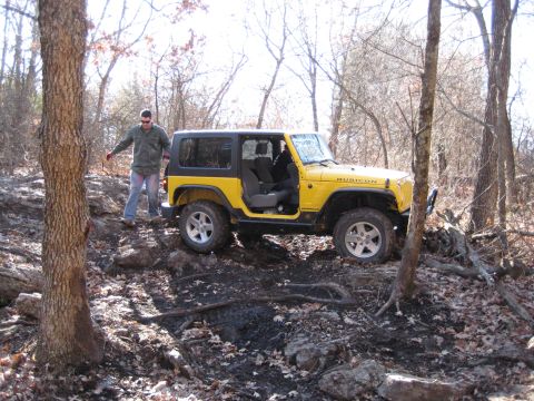 2005 Jeep Wrangler - A yellow Jeep Wrangler Rubicon navigates a rough, wooded terrain with a person standing nearby.