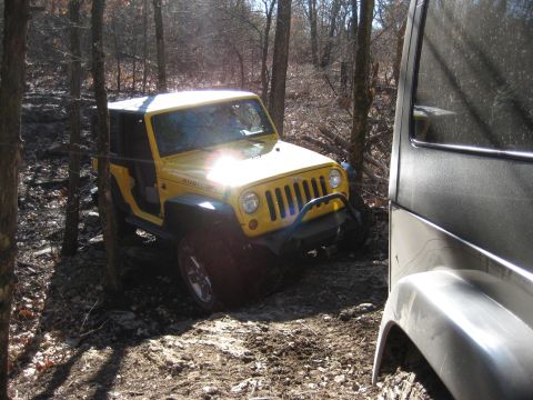 2005 Jeep Wrangler - A yellow Jeep Wrangler navigates a muddy, wooded path, with another vehicle partially visible on the right.