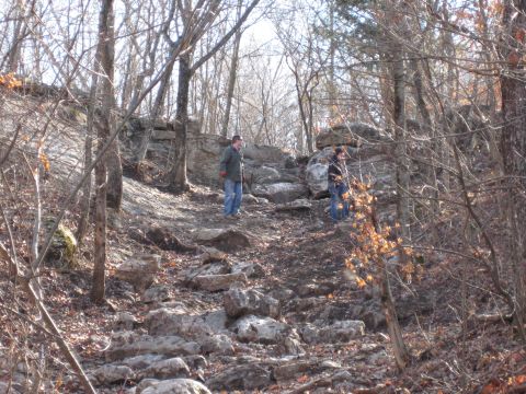 2005 Jeep Wrangler - A scene of two individuals navigating a challenging, rocky terrain in a wooded area during daylight.