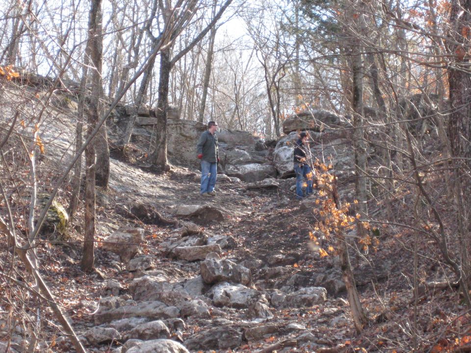 Two people stand on a rocky, wooded trail with bare trees and large boulders