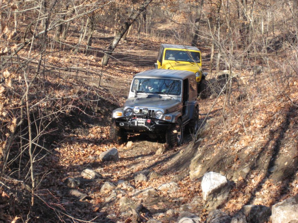 Two Jeep Wranglers driving on a rocky, wooded trail in autumn. The front Jeep is silver, the rear is yellow
