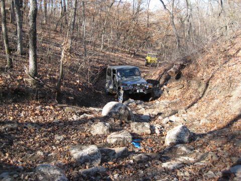 2005 Jeep Wrangler - Two Jeeps navigate a challenging, rocky path through a wooded area during an off-road excursion.