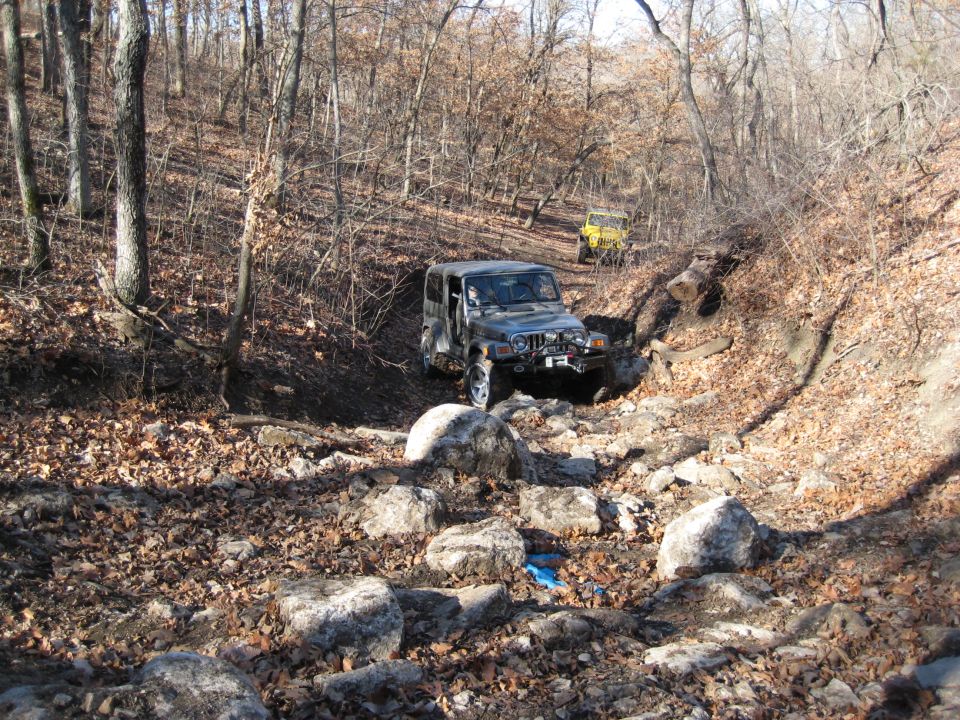 Two Jeeps driving on a rocky, wooded trail. The front Jeep is dark colored and the one behind it is yellow