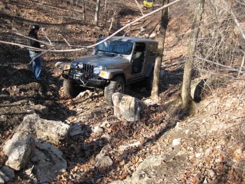 2005 Jeep Wrangler - A gray Jeep Wrangler tackles a challenging rocky trail in a wooded area, with another vehicle visible in the distance.