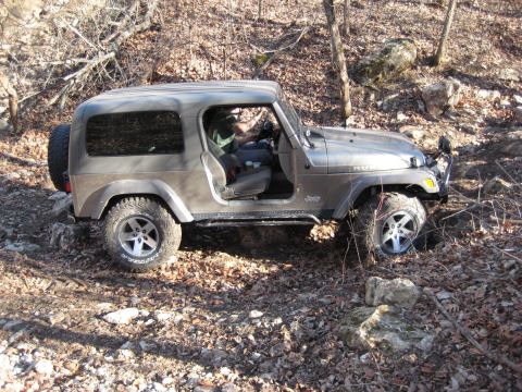 2005 Jeep Wrangler - A gray 2005 Jeep Wrangler navigates a rough, wooded path, its driver's door ajar.