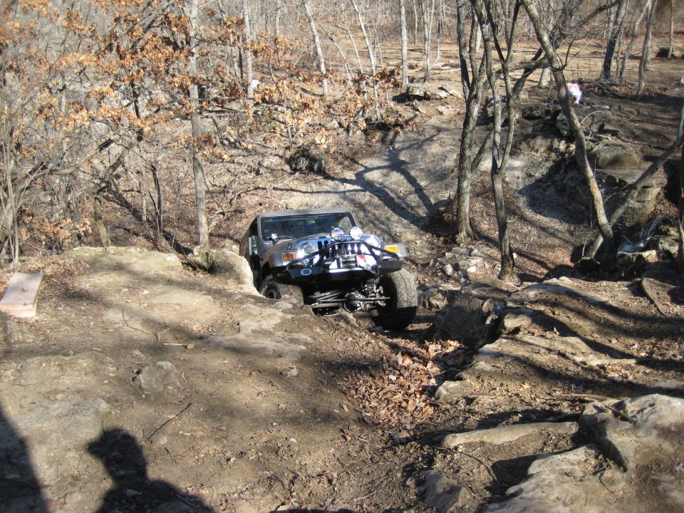 Jeep Wrangler navigating a rocky, wooded trail. The vehicle is angled, with its front wheels turned