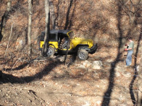 2005 Jeep Wrangler - A yellow 2005 Jeep Wrangler is shown off-roading through a wooded area with rocky obstacles.