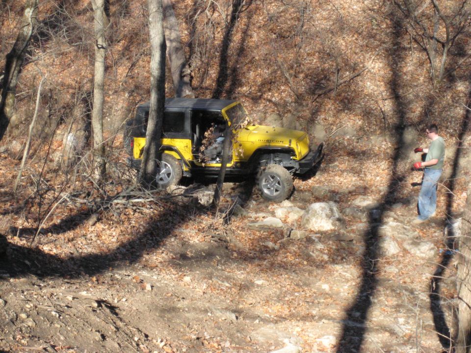 Yellow 2005 Jeep Wrangler navigating a rocky, wooded trail with a person standing nearby