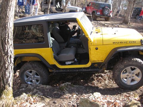 2005 Jeep Wrangler - A yellow Jeep Wrangler navigates a challenging off-road course, with other vehicles visible in the wooded area.