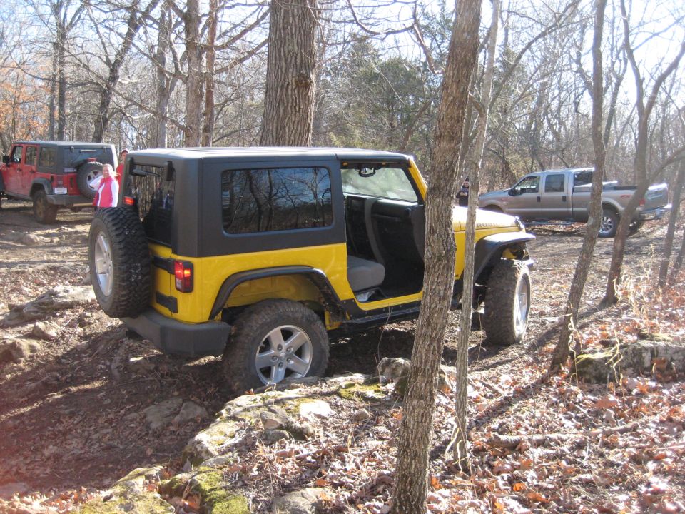 Yellow Jeep Wrangler with black top, door open, on a rocky trail with a red Jeep and a pickup truck in the background