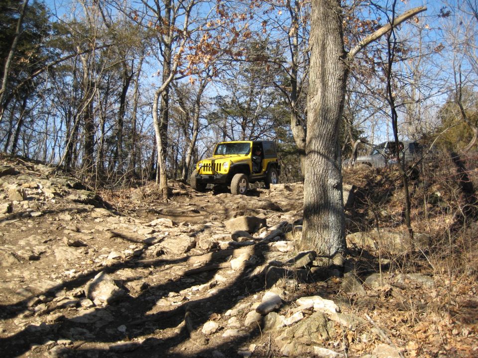 Yellow Jeep Wrangler on a rocky, wooded trail, with another vehicle behind it