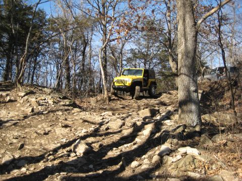 2005 Jeep Wrangler - Yellow Jeep Wrangler driving on a rocky trail in a wooded area.