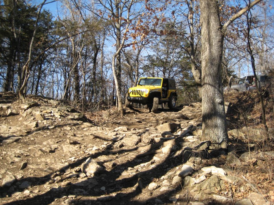 Yellow Jeep Wrangler navigating a rocky off-road trail with another vehicle visible in the background