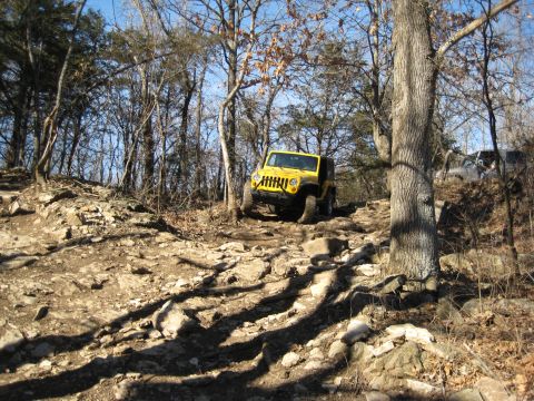 2005 Jeep Wrangler - A yellow Jeep Wrangler navigates a challenging, rocky off-road trail with trees and sparse foliage.