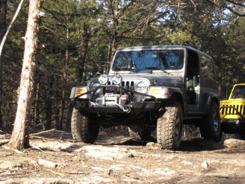 2005 Jeep Wrangler - A gray Jeep Wrangler navigates a wooded, rocky terrain, with another Jeep partially visible in the background.