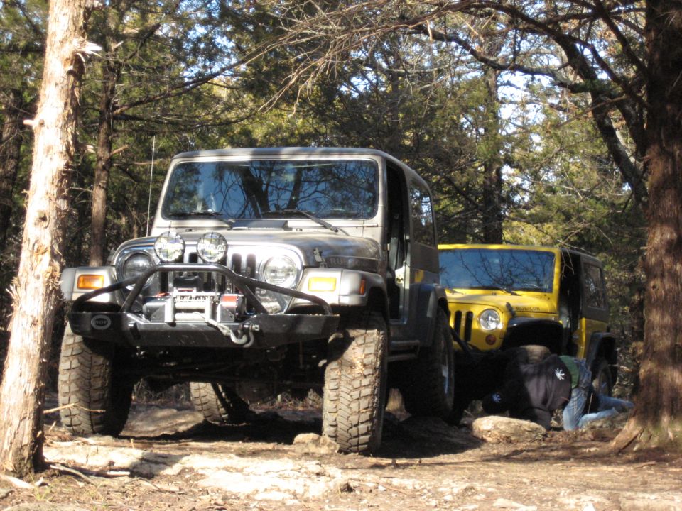 Two Jeep Wranglers parked on a dirt trail with trees around. The front Jeep is gray, the rear is yellow