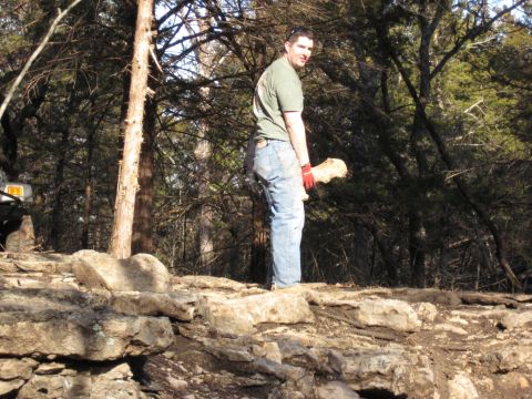 2005 Jeep Wrangler - A man stands in a rocky, wooded area holding a large piece of wood, with a portion of a vehicle visible on the left side of the frame.