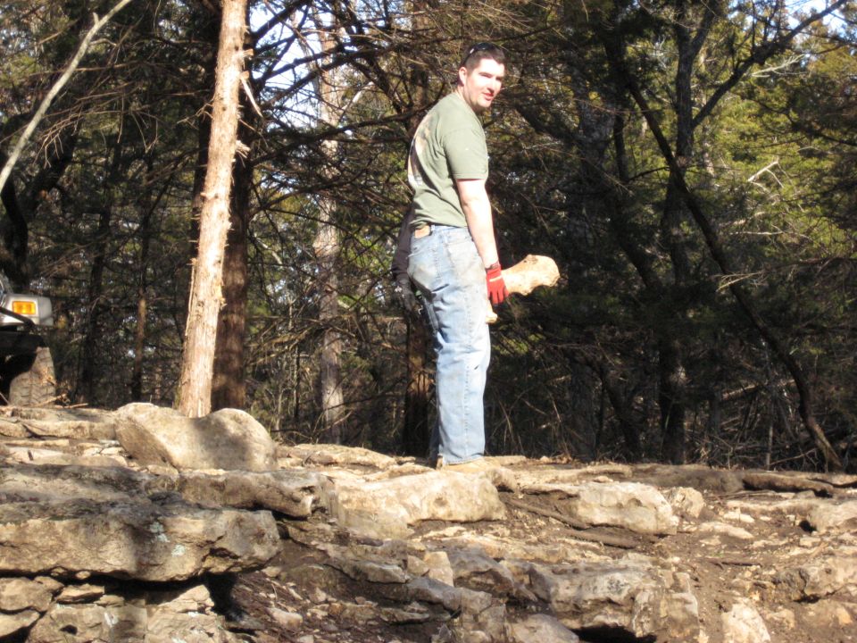 Man holding wood in a wooded area with a partial vehicle visible on the left