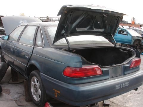 1993 Ford Taurus - Rear view of a 1993 Ford Taurus SHO at a junkyard, showing the open trunk and other vehicles in the background.